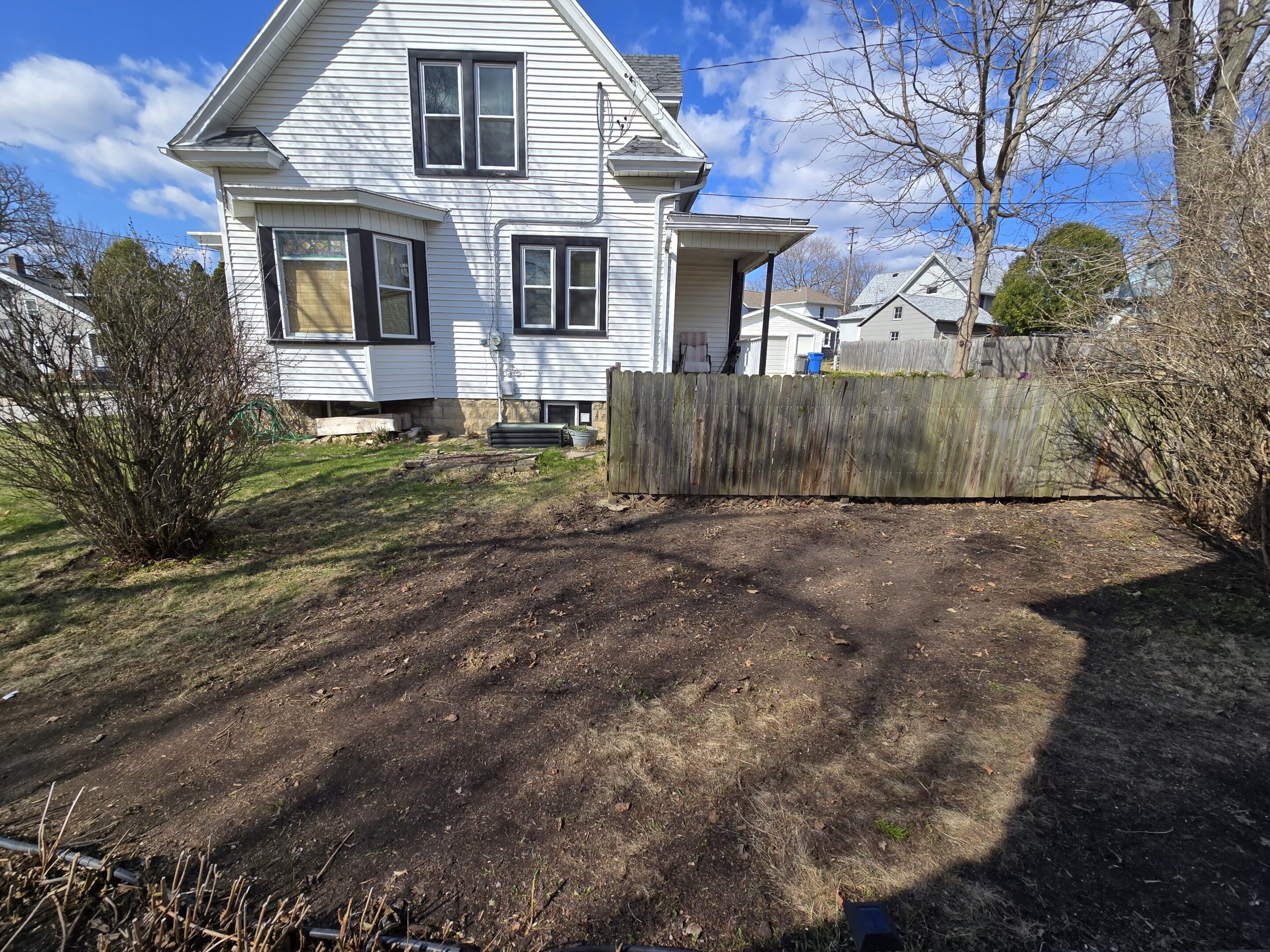 Freshly tilled garden soil in a residential backyard under a partly cloudy sky.