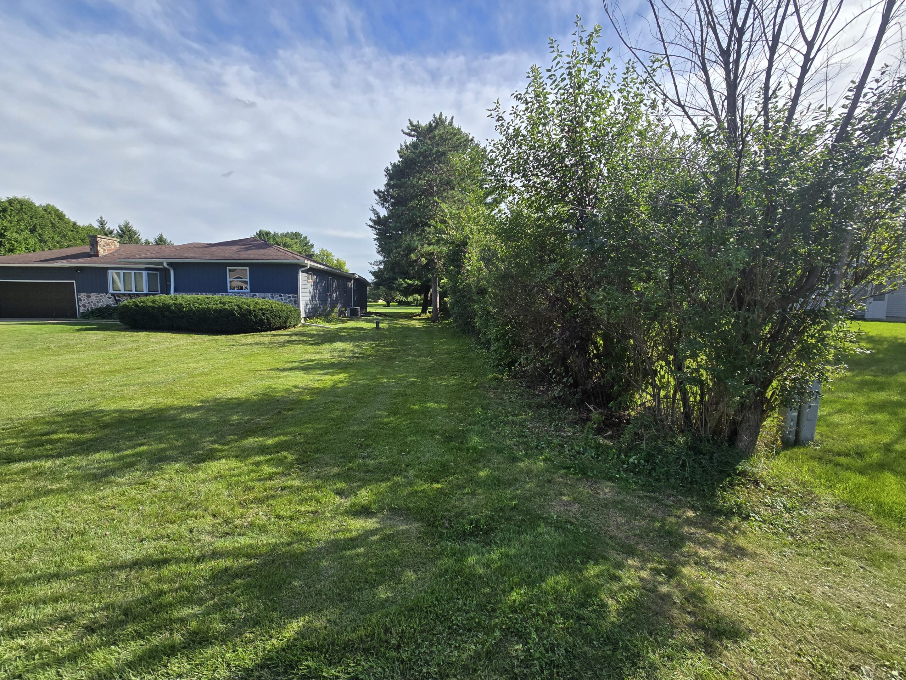 Green lawn with a row of trees and a house in the background under a partly cloudy sky.
