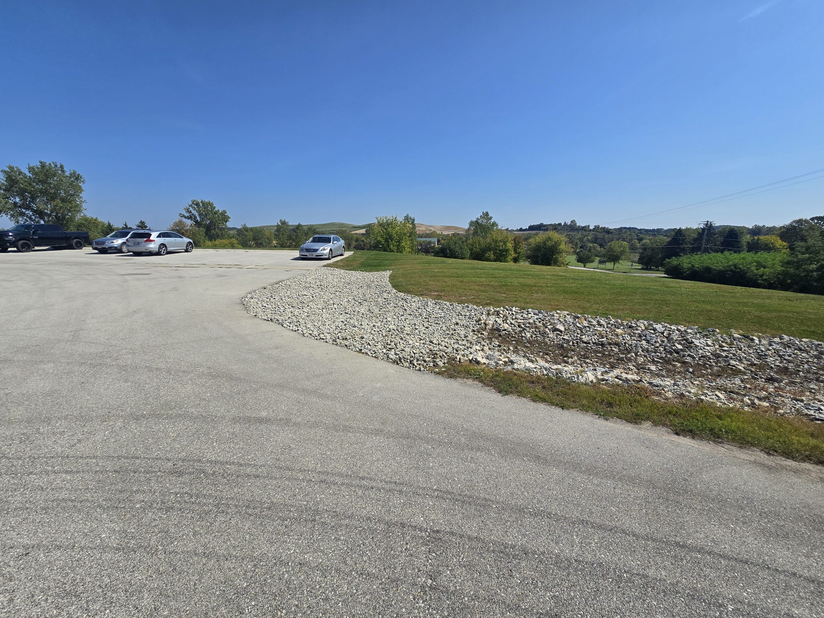 A quiet rural road with parked cars and green fields under a clear blue sky.