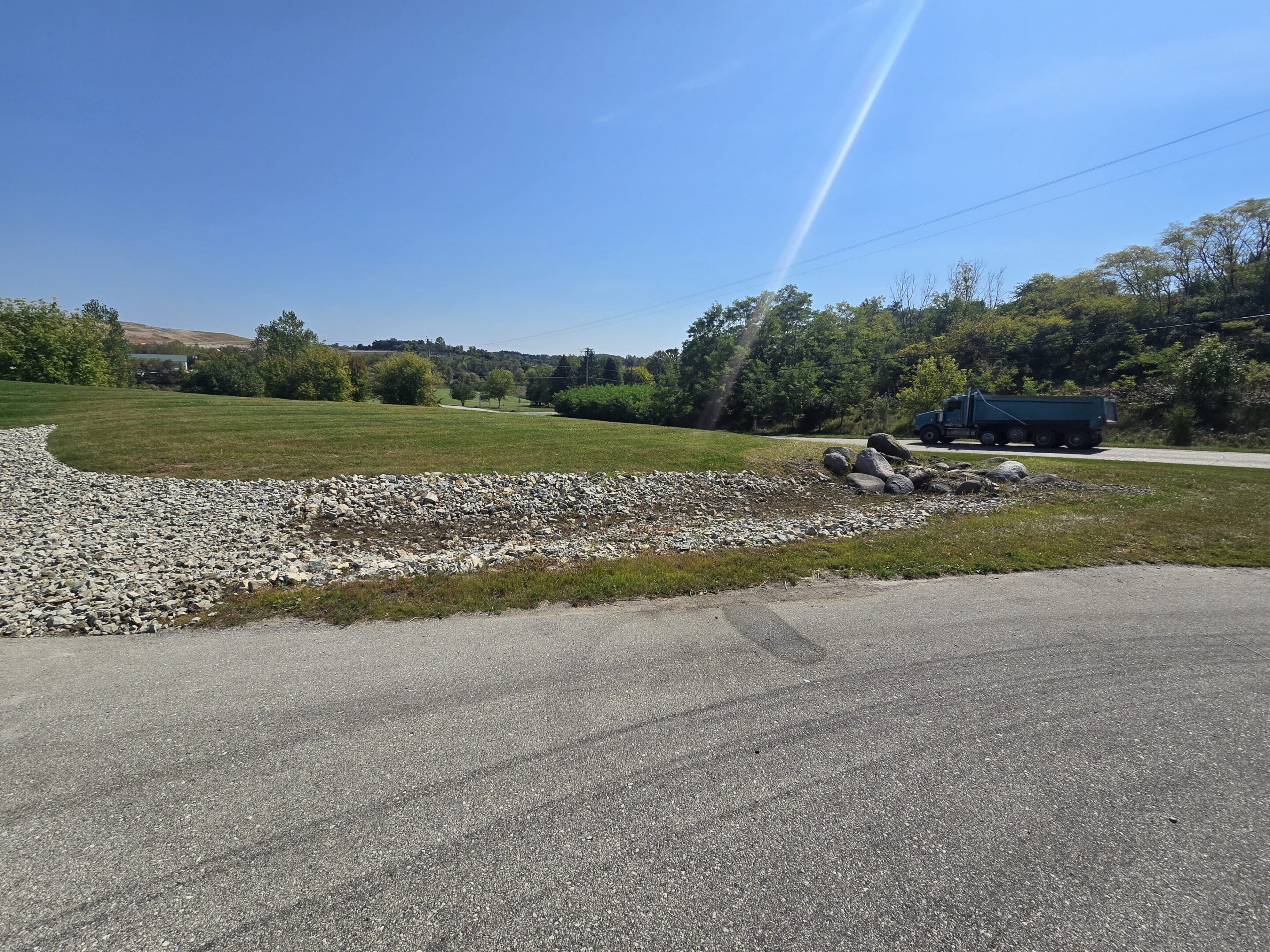 A rural roadside scene with a stone wall and greenery under a clear blue sky.